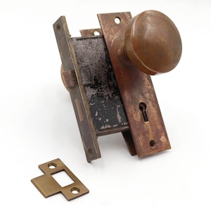 Peut inclure: Ensemble de bouton de porte et serrure en bronze antique. L'ensemble comprend un bouton rond, une plaque frontale rectangulaire avec un trou de serrure et une gâche séparée. Le métal a un aspect vieilli, suggérant l'âge et l'utilisation.