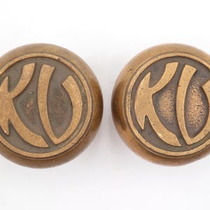 Puede incluir: Dos botones antiguos de color bronce. Cada botón es redondo con un diseño en relieve de las letras "KU" en una fuente estilizada dentro de un borde circular. Los botones tienen un aspecto vintage y envejecido.
