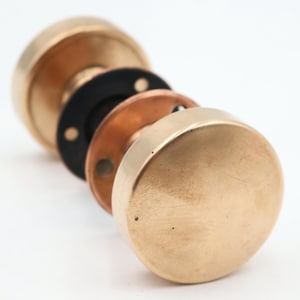 Könnte beinhalten: Nahaufnahme eines zerlegten antiken Messing-Türknaufs. Die Komponenten umfassen einen großen, runden Knopf, eine schwarze Unterlegscheibe, eine kupferfarbene Platte und einen kleineren Knopf. Die Hardware ist in einer Reihe angeordnet und zeigt ihre Konstruktion.