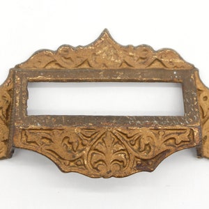 May include: Antique gold-toned metal drawer pull with an ornate design and a rectangular opening. The pull has a decorative floral pattern and two small holes on either side.