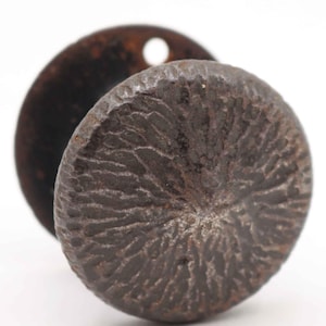 May include: Close-up of a dark brown, textured metal knob with a circular design. A smaller, rusty brown metal disc with a hole is behind the knob. The knob has a rough, organic appearance.