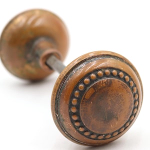 May include: Antique bronze door knob with a round, decorative face featuring a dotted border. The knob has a weathered appearance, suggesting age and use. The hardware is mounted on a metal rod.