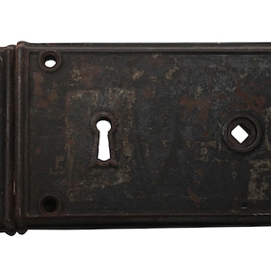 May include: A rusty metal door lock with a keyhole and a square-shaped hole. The lock has a dark brown patina and is rectangular in shape.