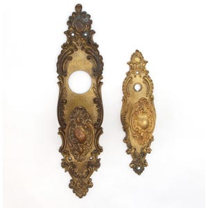 May include: Two ornate brass door plates with intricate designs. The larger plate has a keyhole cutout and a smaller decorative element in the center. The smaller plate has a single decorative element in the center.