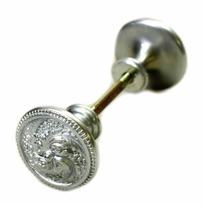 May include: A silver door knob featuring an embossed floral pattern on the front. The knob has a rounded design and is attached to a cylindrical rod and a smaller, rounded back plate. The hardware is set against a white backdrop.
