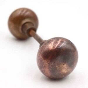 Op de afbeelding: Antieke bronzen deurknop met een rond, bolvormig ontwerp. De knop heeft een verweerde uitstraling met zichtbare patina, wat duidt op leeftijd en gebruik. De verbindingsstang is ook van brons, tegen een witte achtergrond.