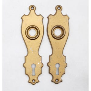 Puede incluir: Dos placas de puerta doradas antiguas con una cerradura y un agujero redondo en el centro. Las placas tienen un diseño decorativo alrededor de los bordes.