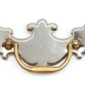 May include: A silver and brass cabinet pull with a decorative, ornate design. The pull has a curved, D-shaped handle.
