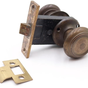 May include: An antique door lock set with a brass finish. The set includes a black metal lock body, two round doorknobs, a strike plate, and a rectangular faceplate. The hardware is likely vintage and shows signs of wear.