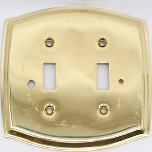 May include: A polished brass double light switch plate with a decorative border. The plate has two rectangular cutouts for light switches and four screw holes.