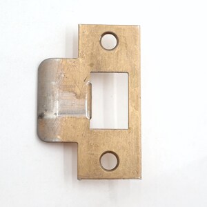 May include: A brass-colored door strike plate with a rectangular opening and two circular holes. The plate has a curved edge on one side and is designed for door hardware installation. The metal has a slightly worn appearance.