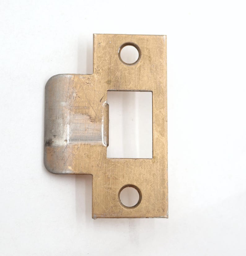 May include: A brass-colored door strike plate with a rectangular opening and two circular holes. The plate has a curved edge on one side and is designed for door hardware installation. The metal has a slightly worn appearance.