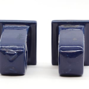 May include: Two blue ceramic wall mounted candle holders. The holders are square shaped with a rounded top and a small lip around the top edge.