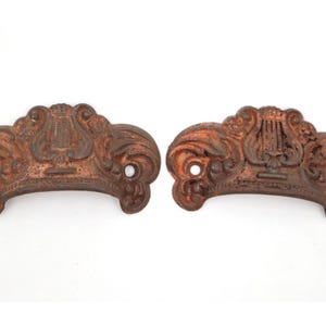 May include: Two antique bronze-colored drawer pulls. Each pull features a decorative lyre design in the center, with ornate scrollwork on either side. The pulls have two holes for mounting and a curved handle shape.