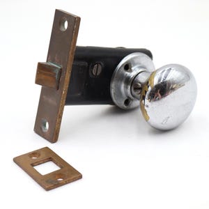 May include: An antique door knob set with a chrome finish and a black body. The set includes a rectangular strike plate and a separate rectangular plate. The metal has a tarnished appearance.