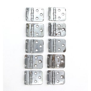 Puede incluir: Diez bisagras de metal plateado, cada una con cuatro agujeros, dispuestas en dos filas de cinco. Las bisagras están ligeramente desgastadas y tienen un aspecto rústico.