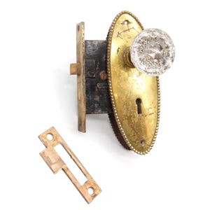 May include: Antique brass door knob with a clear glass crystal knob and a keyhole. The knob is oval shaped with a beaded edge. The door knob is attached to a brass plate with a black metal latch.