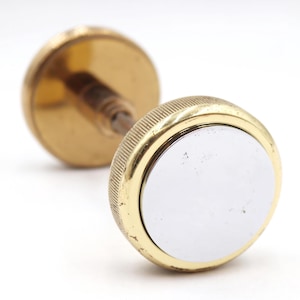 May include: A vintage dumbbell with a gold finish. It has a ridged handle and two circular weights, one with a silver centre. The dumbbell is set against a white background.