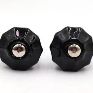 Può includere: Due pomelli neri ottagonali per mobili con finitura lucida e centro argentato. Questi pomelli decorativi sono probabilmente utilizzati per mobili come cassetti o armadi. I pomelli sono su uno sfondo bianco.