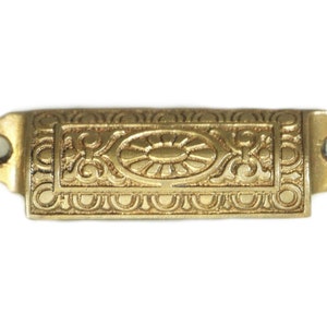 May include: A gold-toned metal drawer pull with an ornate design. The pull has a rectangular shape with a raised oval in the center. The oval has a sunburst design.