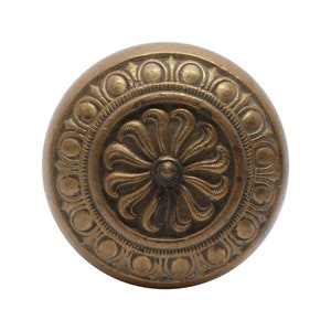 Può includere: Una maniglia per porta rotonda in ottone con un motivo floreale decorativo al centro. La maniglia ha un bordo circolare rialzato con un motivo ripetitivo di piccoli cerchi.