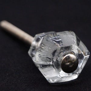 Puede incluir: Un pomo de armario de vidrio transparente con un inserto de metal plateado. El pomo tiene un diseño facetado y está unido a un tornillo de metal.