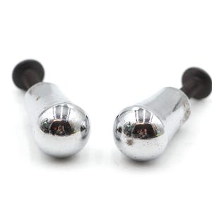 May include: Two chrome-plated cabinet knobs with a rounded, bulbous design. Each knob has a dark, cylindrical stem and a circular, black end. The knobs are set against a plain white background.
