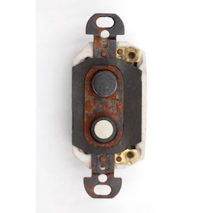 May include: A vintage electrical switch with a rectangular black faceplate, two circular buttons, and a rusty metal frame. The switch has brass connectors and a white ceramic base, set against a white background.