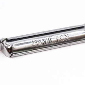May include: A silver metal drawer pull with the word "HAMILTON" embossed on the front.