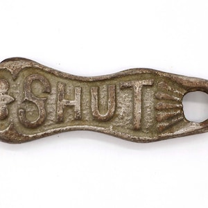 Peut inclure: Un loquet de porte en fonte avec un trou pour une poignée. Le loquet est marqué "SHUT" en lettres en relief.