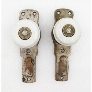 May include: Two door knobs with white ceramic centers and metallic bases. Each knob features a circular metal plate in the center with a cross pattern. The rectangular metal backplates show signs of wear and have two screw holes for mounting.