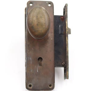 May include: An antique door lock set with a tarnished brass finish. The set includes an oval-shaped doorknob, a keyhole, and a rectangular backplate. The lock mechanism is visible on the side. The hardware shows signs of age and wear.