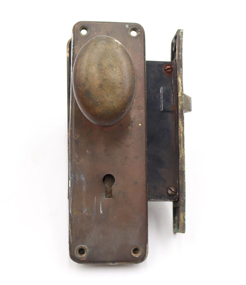 May include: An antique door lock set with a tarnished brass finish. The set includes an oval-shaped doorknob, a keyhole, and a rectangular backplate. The lock mechanism is visible on the side. The hardware shows signs of age and wear.