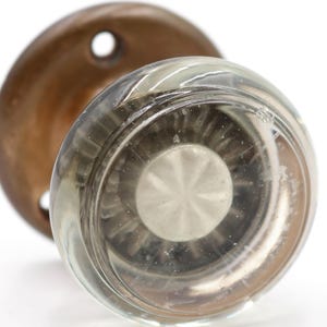 May include: A clear glass doorknob with a circular, ridged design and a metal center. The doorknob is paired with a round, bronze-colored backplate with a single hole. The hardware is set against a white background.