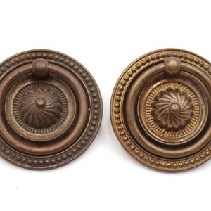 May include: Two antique bronze-colored drawer pulls. Each pull is circular with a central design and a ring handle. The outer edge features a beaded border. The pulls are likely for furniture restoration or decorative use.