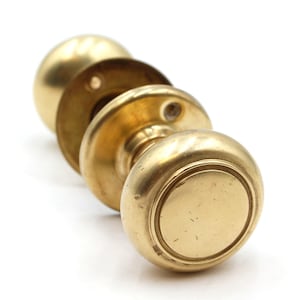 May include: A pair of polished brass door knobs featuring a classic round shape with a smooth, reflective surface. Each knob is attached to a circular mounting plate with visible screw holes, showcasing a metallic gold finish and a simple design.