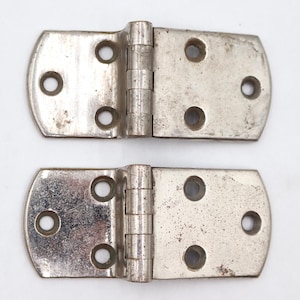 May include: Two vintage, silver-toned metal hinges. Each hinge has a rectangular shape with rounded edges and multiple screw holes. The hinges are connected by a central pin, showing signs of wear and age.