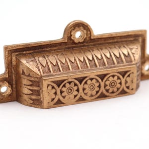 Może przedstawiać: Mosiężny uchwyt do szuflady z misternymi wzorami kwiatowymi i geometrycznymi. Prostokątny uchwyt posiada szereg stylizowanych wzorów kwiatowych i dekoracyjną górę z pojedynczym otworem do montażu. Okucia przeznaczone są do mebli.