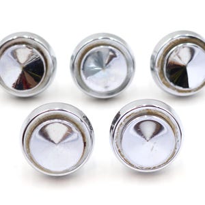 Può includere: Cinque pomelli cromati lucidi con un design circolare. Ogni pomello ha un centro conico riflettente e un bordo arrotondato. I pomelli sono disposti su uno sfondo bianco, mettendo in risalto il loro aspetto lucido.