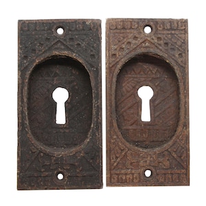 Może przedstawiać: Dwa antyczne, żeliwne osłony otworów na klucz z ozdobnymi wzorami. Osłony są prostokątne z otworem na klucz w środku. Osłony są ciemnobrązowe z rustykalnym wykończeniem.