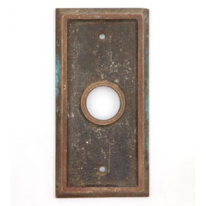 May include: Rectangular antique doorbell button plate with a central circular button and two screw holes. The plate has a dark, aged appearance with a bronze-colored frame and a weathered, textured surface.