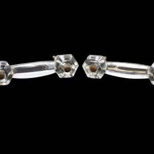 May include: Two clear glass cabinet knobs with a silver metal bar connecting them. The knobs are hexagonal in shape with a small brass screw in the center.