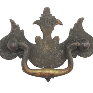 May include: Antique bronze drawer pull with a decorative floral design. The pull features a curved handle and two round knobs on either side. The metal has a distressed finish, giving it a vintage look. Perfect for adding a touch of elegance to furniture.