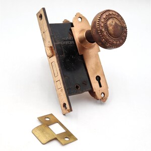 Puede incluir: Un juego de pomo de puerta antiguo con acabado bronce. El juego incluye un pomo redondo decorativo, un mecanismo de cierre rectangular y una placa de cierre separada. El pomo tiene un diseño ornamentado y texturizado.