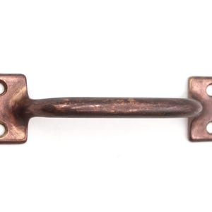 May include: A copper-colored metal handle with a curved bar and rectangular mounting plates. Each plate has two circular holes for screws. The handle is designed for furniture or cabinetry.
