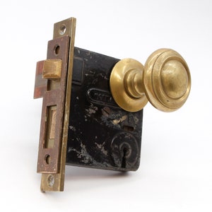 May include: Antique brass door knob and lock set with a black metal plate. The knob has a rounded design with multiple ridges. The lock has a keyhole and a small lever.