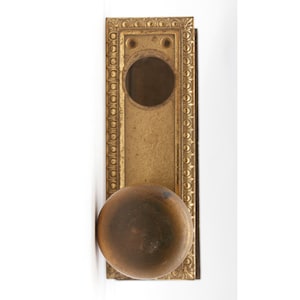 May include: A vintage brass door knob with a decorative frame. The knob is round and has a smooth surface. The frame is rectangular and has a detailed design.