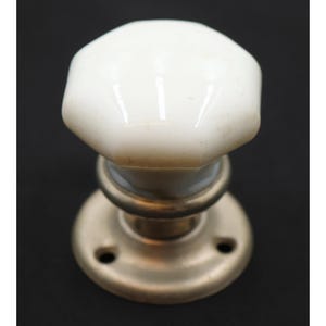 May include: A white octagonal ceramic door knob featuring a smooth, glossy finish. The knob is attached to a circular, brushed metallic base plate that includes two small screw holes for mounting purposes, providing a functional hardware piece.