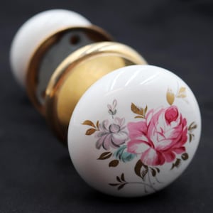 May include: White ceramic door knobs with gold accents. Each knob features a hand-painted floral design with pink and purple roses and gold leaves. The knobs are arranged in a row, showcasing their decorative details.