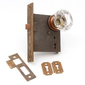 Może przedstawiać: Zestaw antycznych okuć do drzwi z kryształową gałką, ciemną metalową zamkiem i ozdobnymi mosiężnymi płytkami. Zestaw zawiera prostokątną płytkę czołową z ozdobnymi detalami i dwie osłony otworu na klucz.